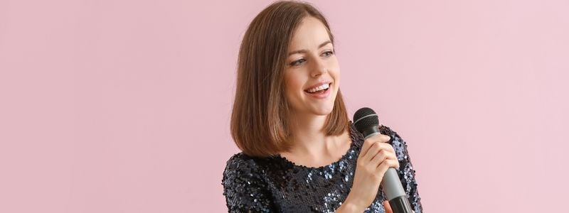 A woman singing with a microphone and light pink background.
